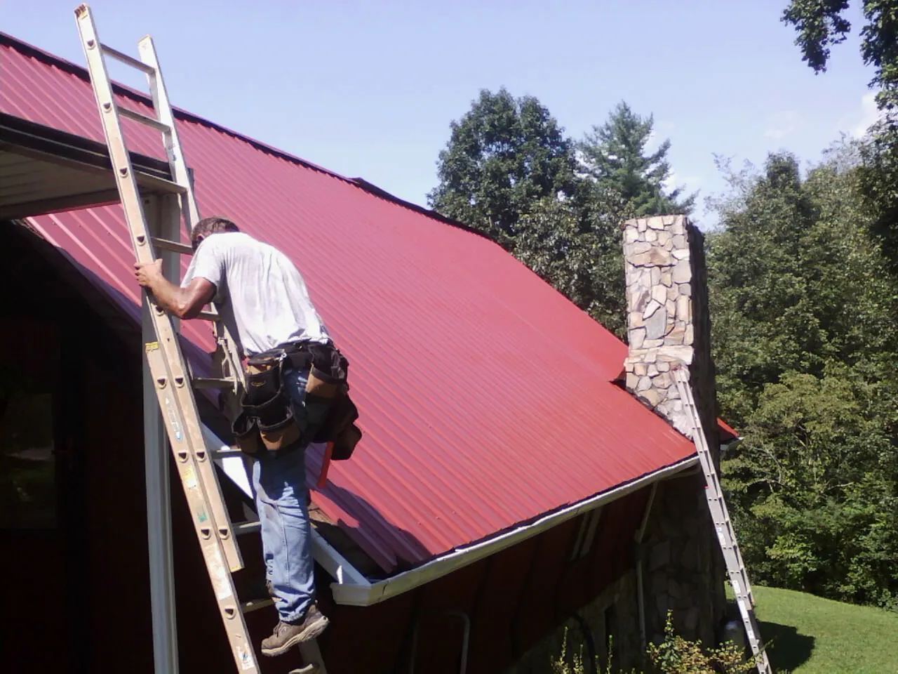 Roofer on a ladder beside a red metal roof and stone chimney — Soffit & Fascia Repair by Roofing Brothers in Society Hill