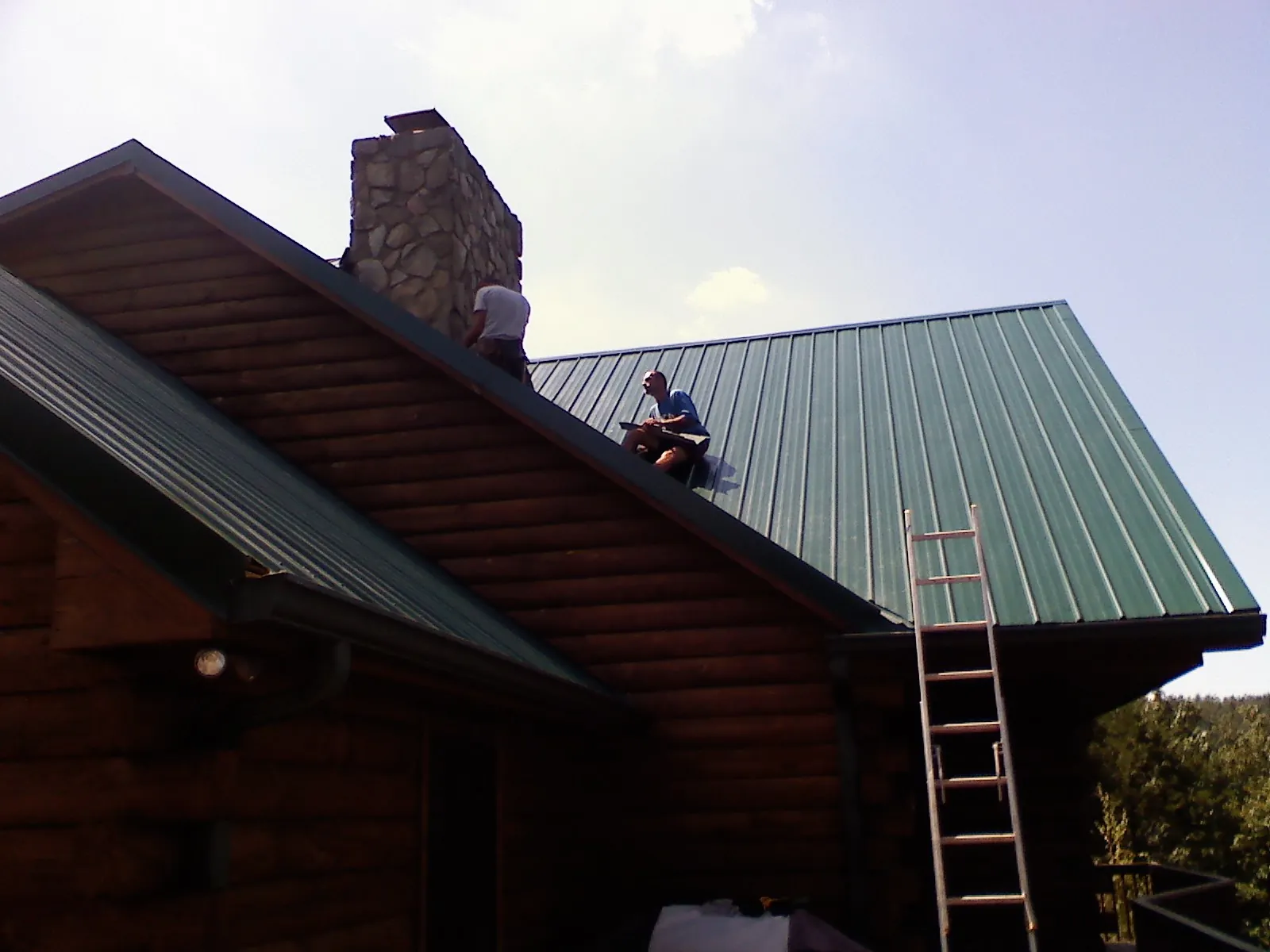 Roofers installing green metal panels beside a stone chimney on a log home in Society Hill